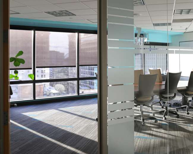 A modern office space features a glass meeting room with a large conference table and chairs, illuminated by natural light from large windows.