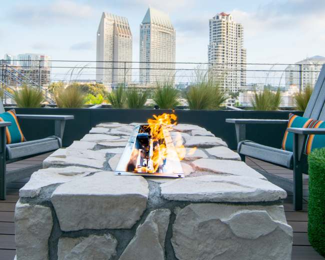 A rooftop patio features a stone fire pit with flames, surrounded by chairs and offering a view of tall buildings against a clear blue sky.