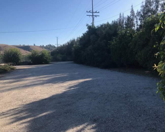 The image shows a gravel parking area bordered by shrubbery and power lines under a clear sky.
