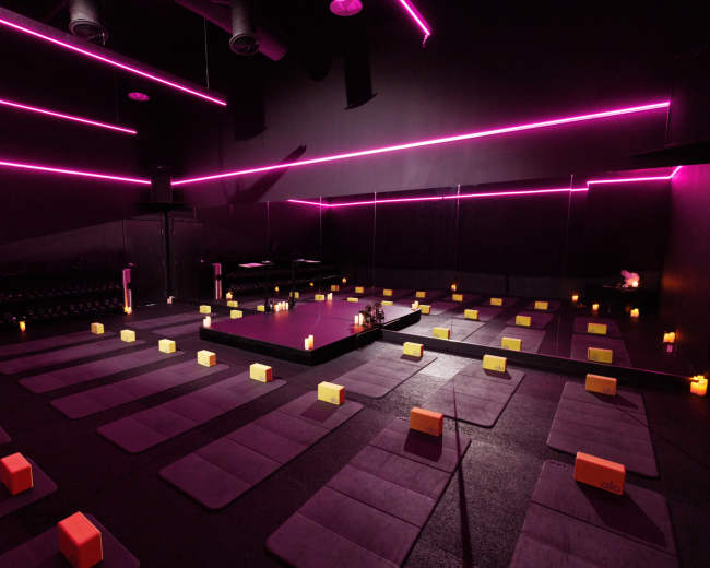 A dimly lit yoga studio features rows of exercise mats and blocks, illuminated by pink neon lights and candles.