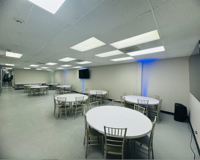 The image shows a spacious room with several round tables surrounded by chairs, featuring a flat-screen TV mounted on one wall and bright overhead lights.