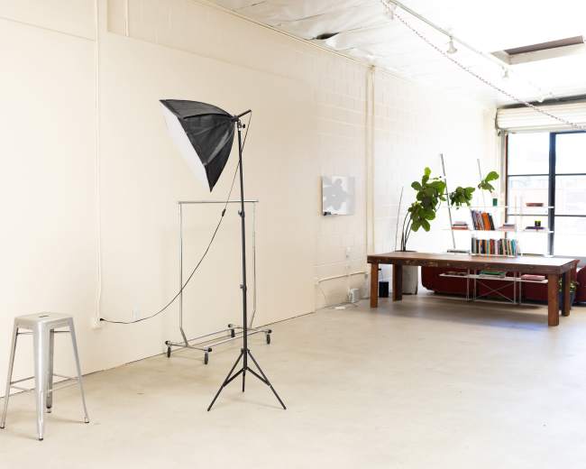 The image shows a bright, minimalistic studio space featuring a photography light on a stand next to a metal stool, with a wooden table and shelving in the background.