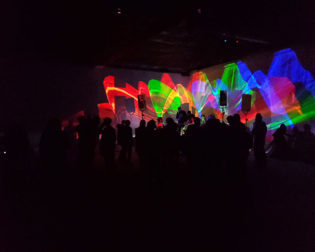 A crowd stands in shadow as colorful light patterns project onto a large wall in a dimly lit venue.