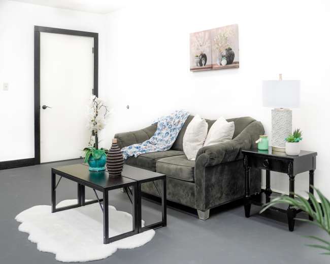 A gray couch with a blue and white throw blanket is placed against a white wall, accompanied by a coffee table and a side table topped with decorative items.