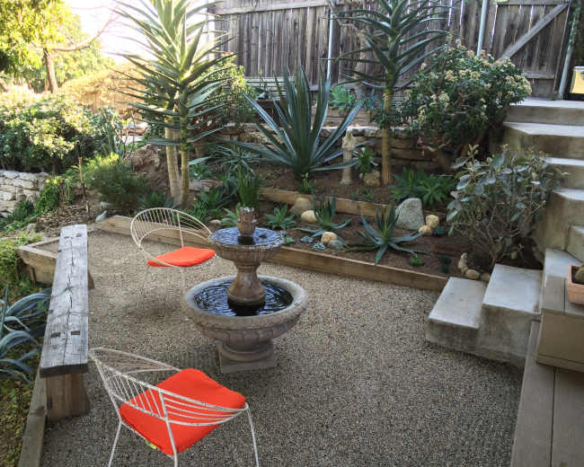 A landscaped garden area featuring a gravel floor, a fountain at the center, two white chairs with orange cushions, and various plants arranged in tiers.