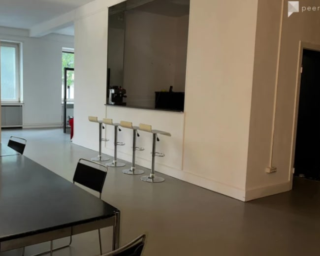 The image shows a modern interior space with a long table, several black chairs, a clothing rack, and a bar area with three high stools.