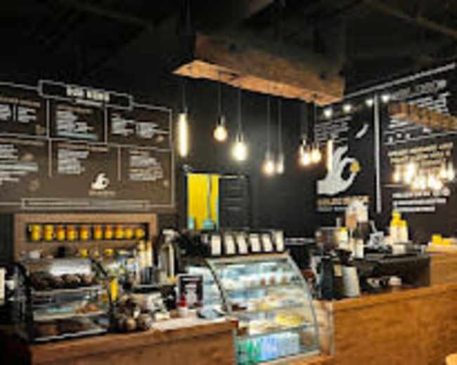 The image shows a coffee shop interior with a wooden counter, display case for food, various menu items on the wall, and hanging light fixtures.