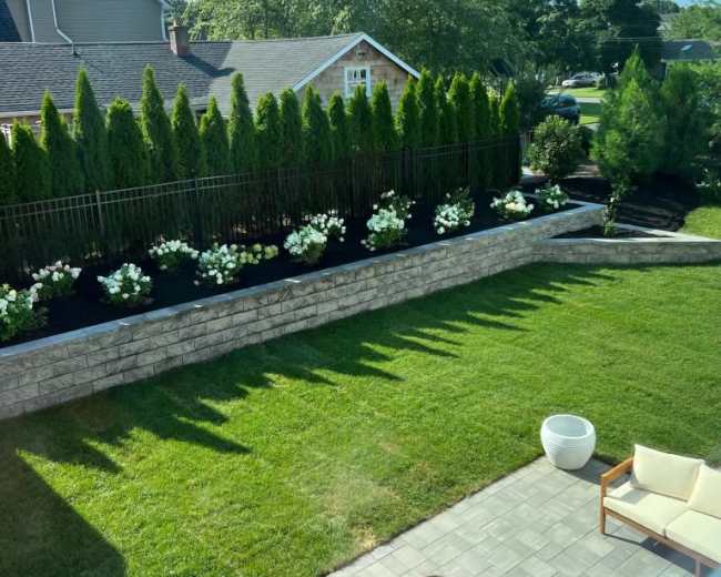 A landscaped backyard features a stone wall with flower beds, green shrubs, and a seating area.