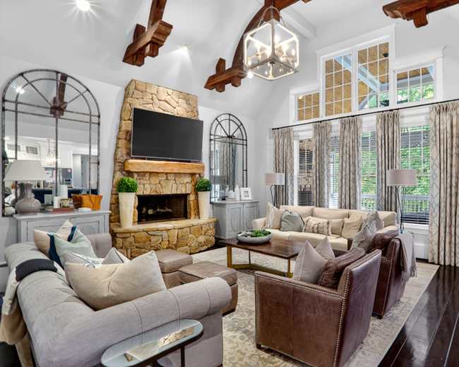 The living room features a stone fireplace, a large flat-screen TV, and a mix of leather and fabric furniture arranged around a central coffee table.