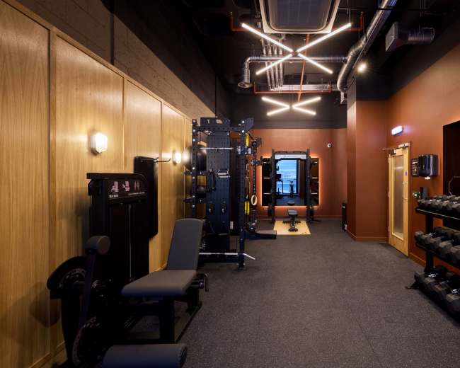 The image shows a modern gym interior featuring weightlifting equipment, dumbbells, and a mirrored wall.