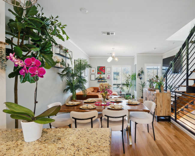 A modern, open-concept living and dining area features a wooden table set for dinner, surrounded by plants and contemporary decor, with a staircase visible in the background.