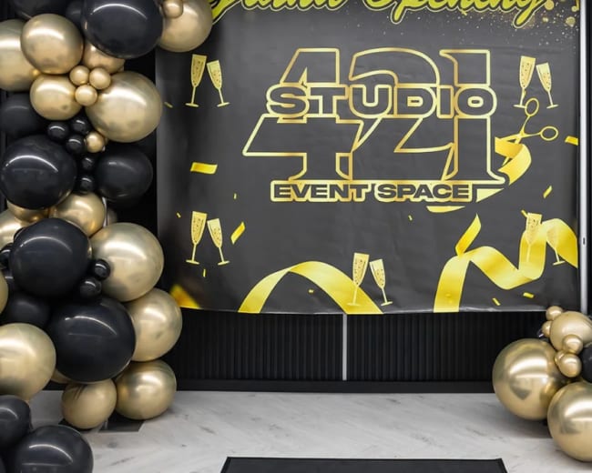 The image shows a grand opening setup for "Studio 421 Event Space," featuring a large black and gold banner, a decorative arrangement of black and gold balloons, and a display of desserts on stands.