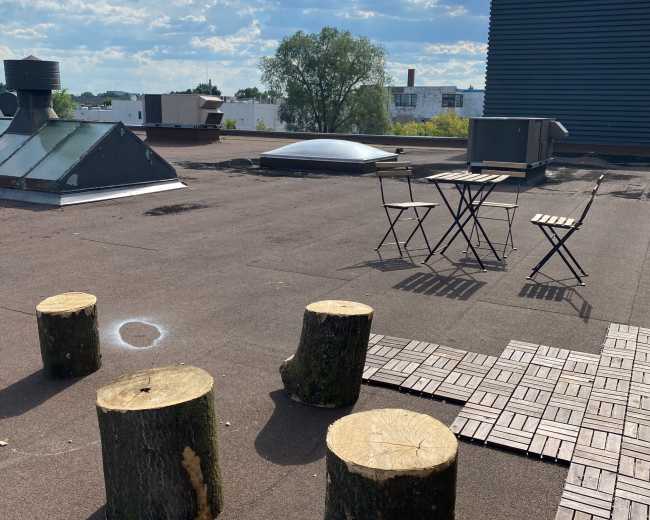 The image shows a rooftop area with wooden stumps arranged as seating, a small table and chairs, and a skylight in the background under a partly cloudy sky.