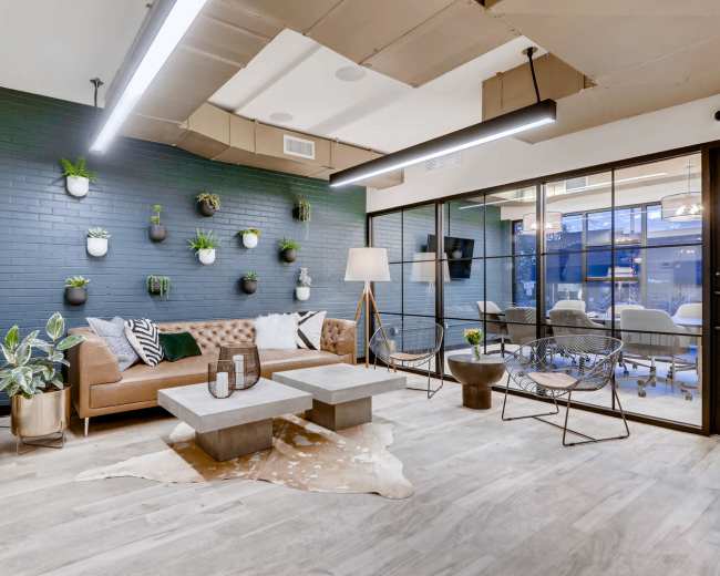 The image shows a modern lounge area featuring a brown sofa, a couple of coffee tables, decorative plants on the wall, and a large glass door leading to a meeting room.