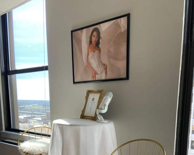 A small table draped with white fabric and adorned with a framed photo and a decorative bust stands between two gold bar stools in a bright room with large windows.
