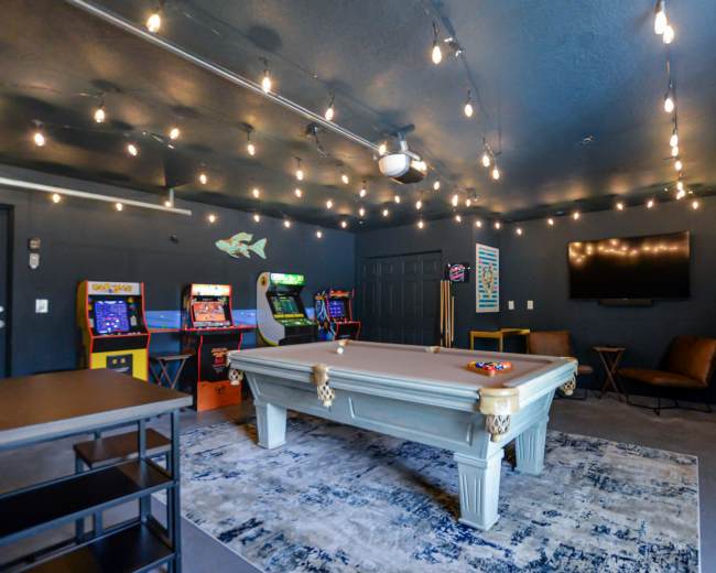 The image shows a game room featuring a billiard table, vintage arcade machines, a television, and string lights overhead, with dark walls and a rug on the floor.