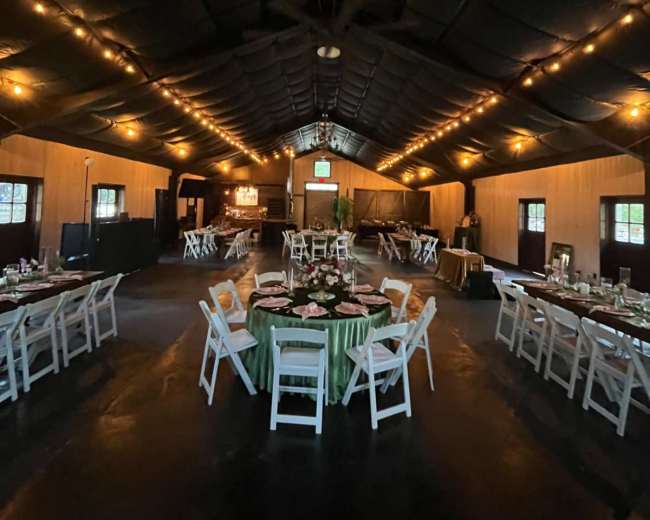 The interior of a large hall features round tables set with white chairs and decorative centerpieces, illuminated by string lights along the ceiling.