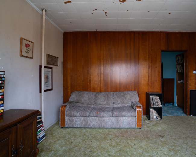 The image shows a sparsely furnished room with a worn gray sofa, wooden paneling on the walls, and a doorway leading to another room.