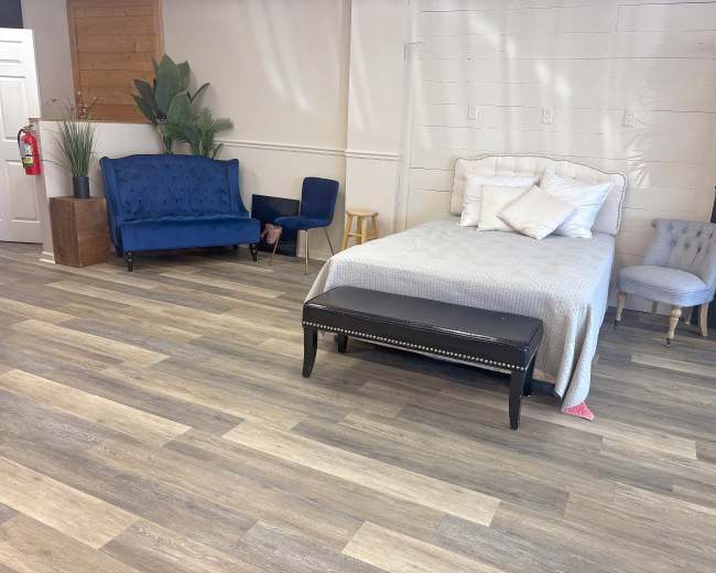 The image shows a spacious room with a bed, a black bench, various seating options, and plants, all arranged on wooden flooring.