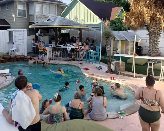 A lively gathering of people is enjoying a pool party in a backyard setting, with some sitting around the pool and others in the water.