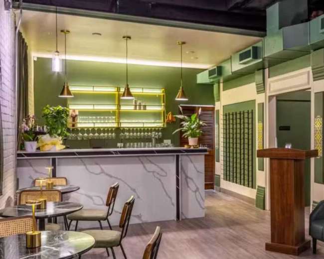 The image shows a stylish bar interior featuring a green color scheme, a marble counter, and a mix of seating arrangements including a leather sofa and small tables.