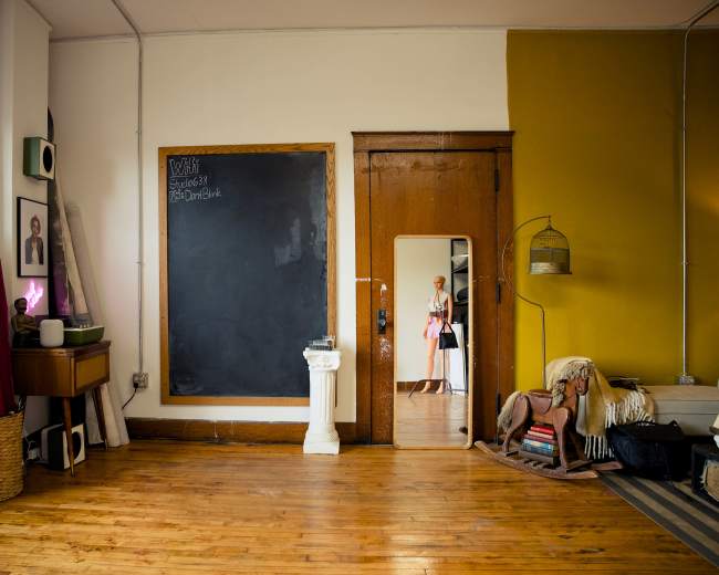 The image shows a spacious room with wooden flooring, featuring a chalkboard on the wall, a mirror, and various decorative items, including a basket and art supplies.