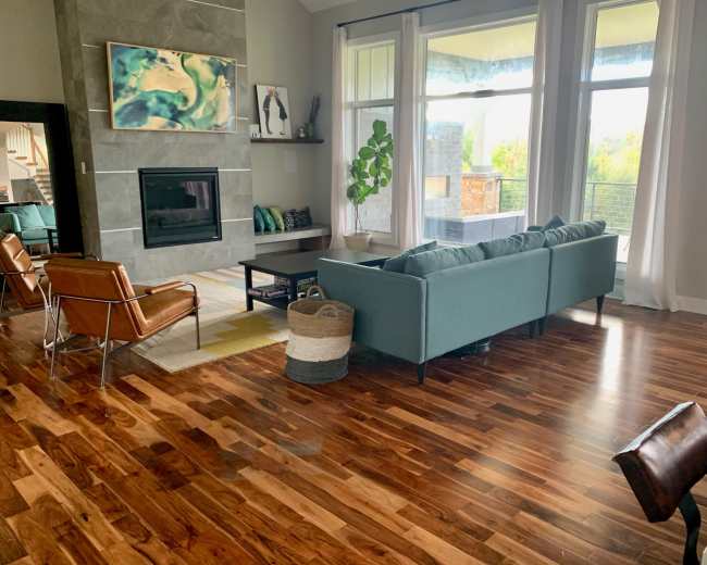A modern living room features a teal sofa, brown leather chairs, a stone fireplace, and large windows overlooking a natural landscape.