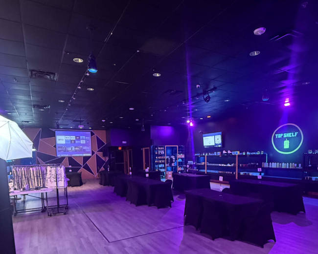The image shows a dimly lit indoor space with tables covered in black cloth, shelves displaying various products, and purple lighting.