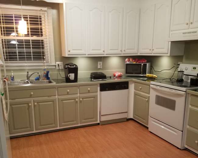 A kitchen with white and light green cabinetry, a stainless steel refrigerator, a white dishwasher, and a stove, all on a wooden floor.