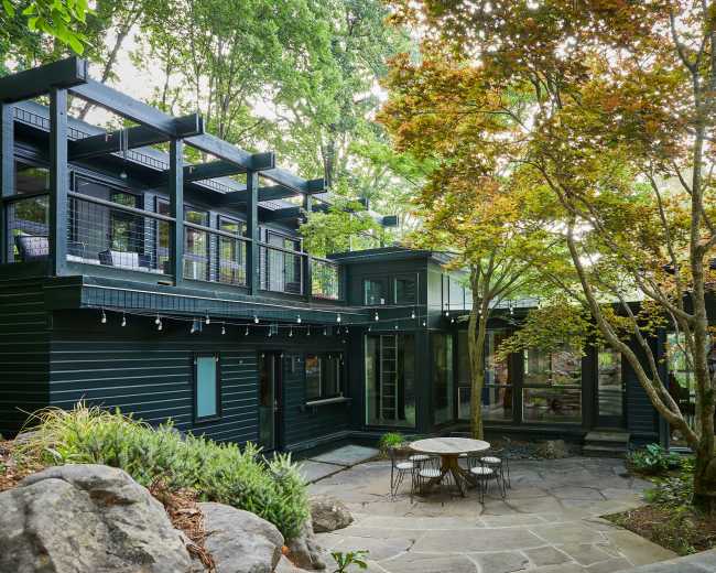 The image shows a modern house with dark exterior walls, surrounded by lush greenery and a stone patio with a circular table.