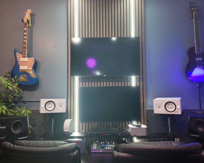 The image shows a modern music production studio featuring two guitars mounted on the wall, a dual-screen setup in the center, and various audio equipment on a desk.