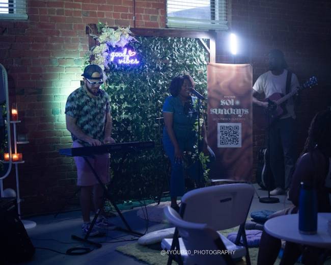A singer performs on stage while accompanied by a keyboardist and a guitarist in a dimly lit venue decorated with greenery.