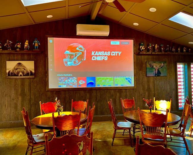 A large screen displaying Kansas City Chiefs highlights hangs on a wall in a dining area with wooden tables and chairs.