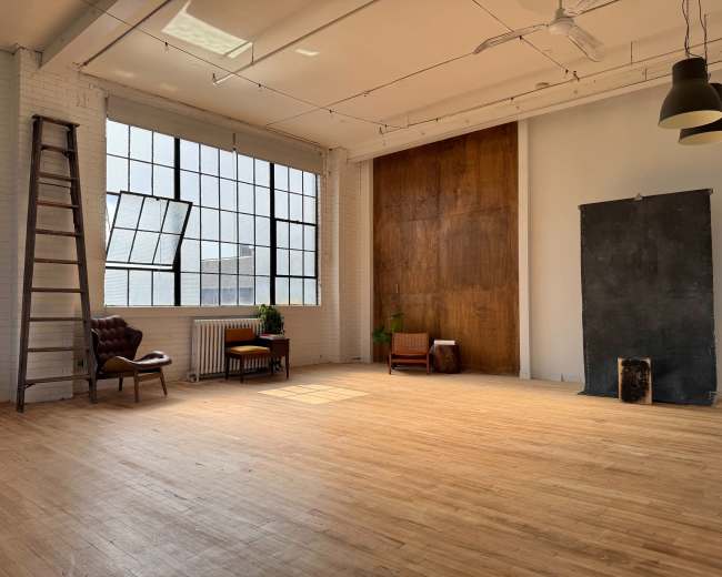 The image depicts a spacious, well-lit studio with a wooden floor, large windows, a ladder, and minimal furnishings including a chair and a small table.