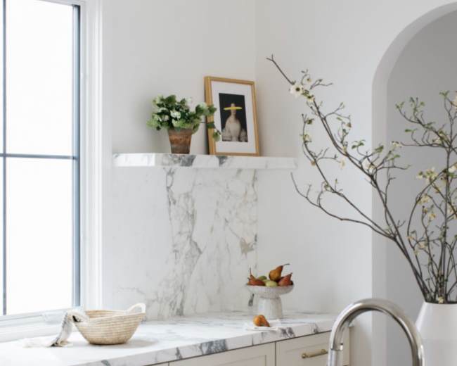 A modern kitchen with marble countertops, a sink, and a window providing natural light, featuring a vase with branches and a framed photo on the wall.