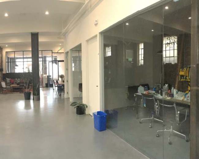 The image shows a spacious, industrial-style office with exposed brick walls, large windows, and both communal and private work areas.