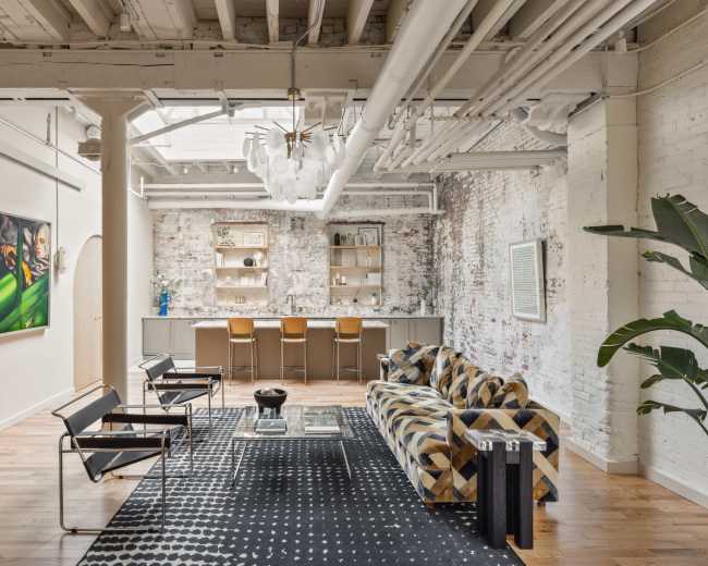 The image depicts a spacious, modern living area with exposed brick walls, wooden flooring, a patterned sofa, and a bar setup in the background.