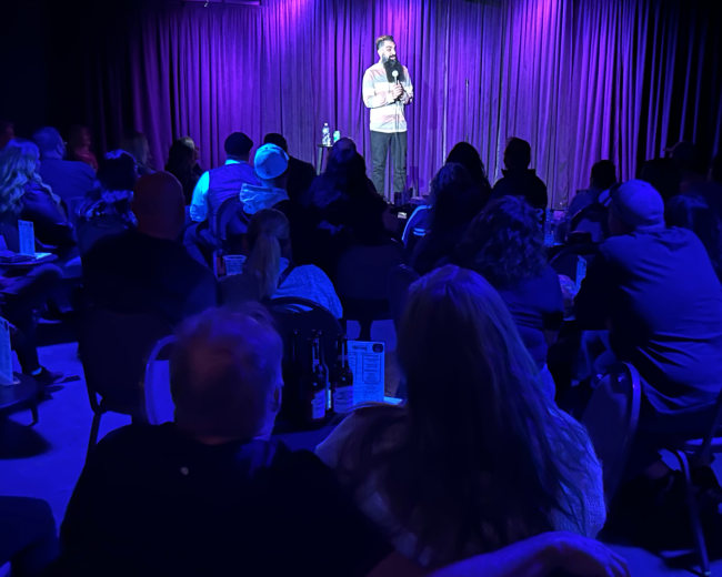A comedian performs on stage in front of an audience seated at tables in a dimly lit venue with purple curtains.