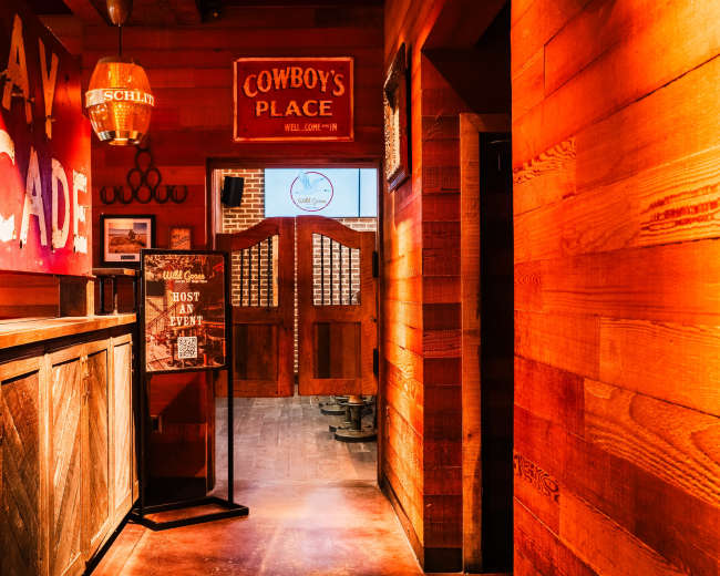 The image shows a wooden hallway with rustic decor, featuring signs for an arcade and a place called "Cowboy's Place," leading to double doors at the end.