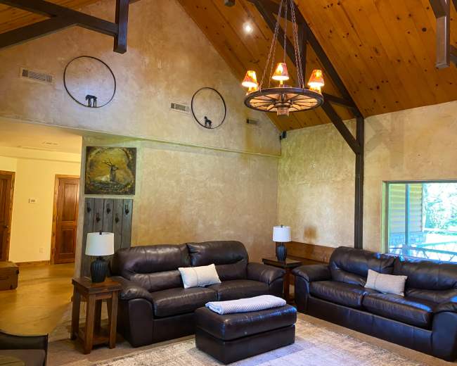 The image features a spacious living room with two dark leather sofas, a central ottoman, and a large window, all set against a backdrop of wooden beams and a textured wall.