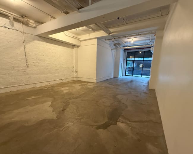 The image shows a spacious, empty room with concrete flooring and white walls, featuring a large window at the far end.