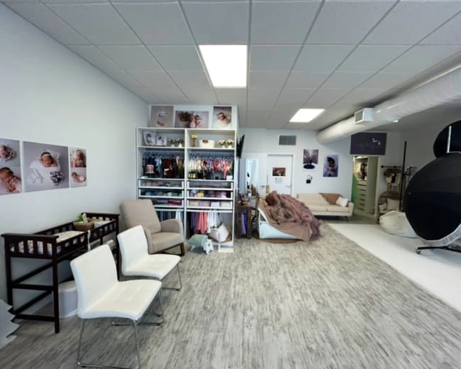 The image shows a photography studio with a seating area, shelving units displaying various items, and a backdrop setup in a bright, open space.