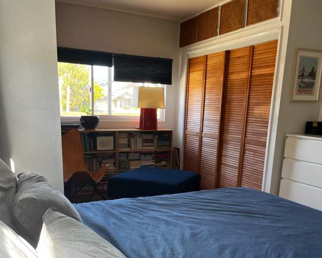 The image shows a bedroom with a bed in the foreground, a bookcase against the wall, and a chair beside a lamp near a window.