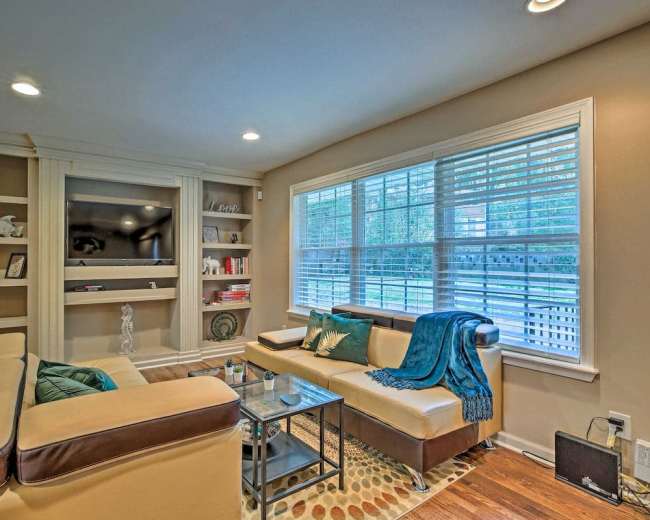 A living room with a beige sofa, a glass coffee table, built-in shelves, and large windows with white blinds.