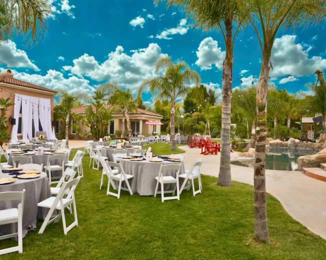The scene shows a spacious outdoor area set up for an event, featuring round tables with gray tablecloths, white chairs, and a pool surrounded by palm trees against a backdrop of blue sky with fluffy clouds.
