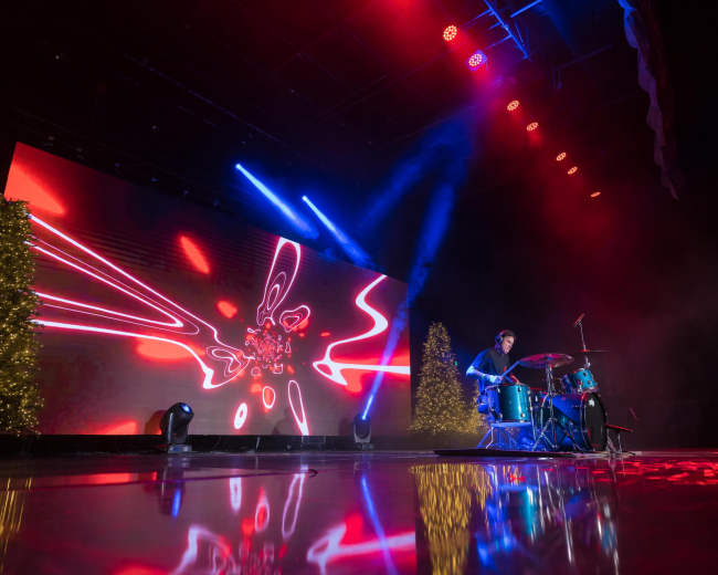 A drummer performs on stage in front of a large illuminated screen with vibrant patterns and two decorated trees.