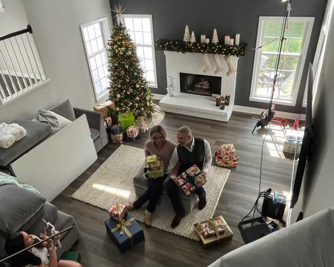A couple sits by a decorated Christmas tree with gifts arranged around them in a cozy living room.
