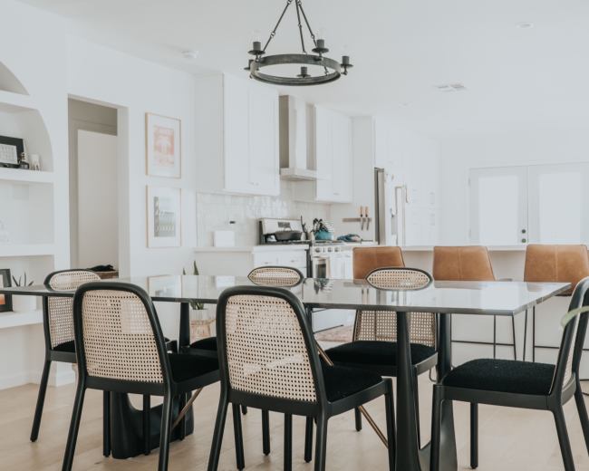 A modern dining area features a glass table surrounded by black and tan chairs, with a kitchen visible in the background.