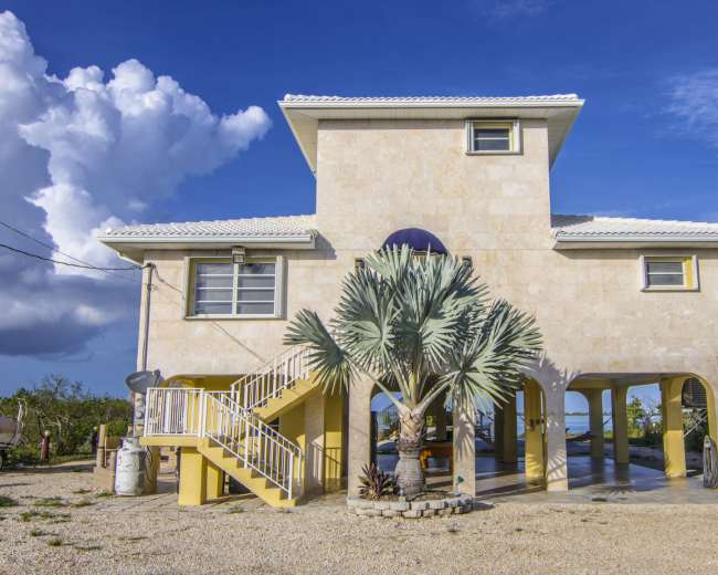 A two-story stone house with a palm tree in front and a staircase leading to the upper floor is set against a blue sky with clouds.