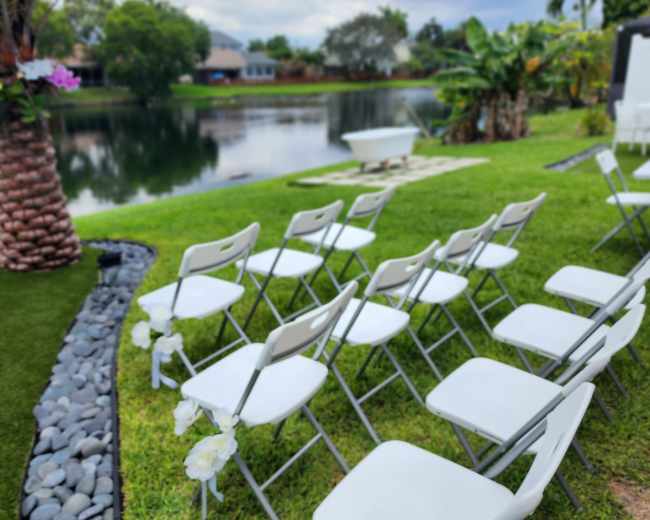 Rows of white chairs are arranged on a grassy area by a calm waterway, flanked by palm trees and lush greenery.